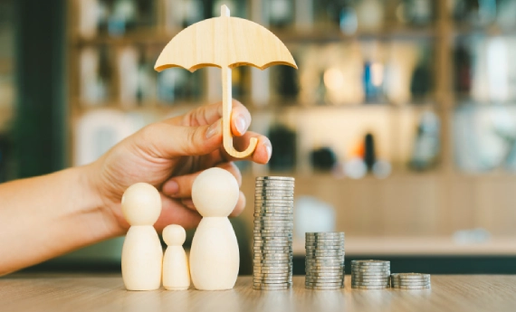 Wooden family figures protected by an umbrella and surrounded by coin stacks.