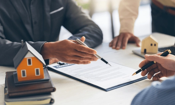 Person signing a document with a small house model nearby.